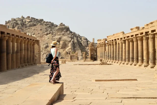 A solo female traveler in a hat and floral skirt explores the ancient ruins of a temple in Egypt, with stone columns and rocky hills in the background.