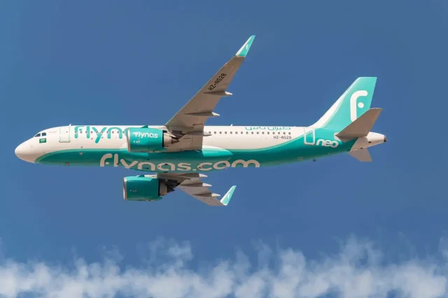 A Flynas Airbus A320neo aircraft in flight, displaying the airline’s turquoise and white livery against a clear blue sky.