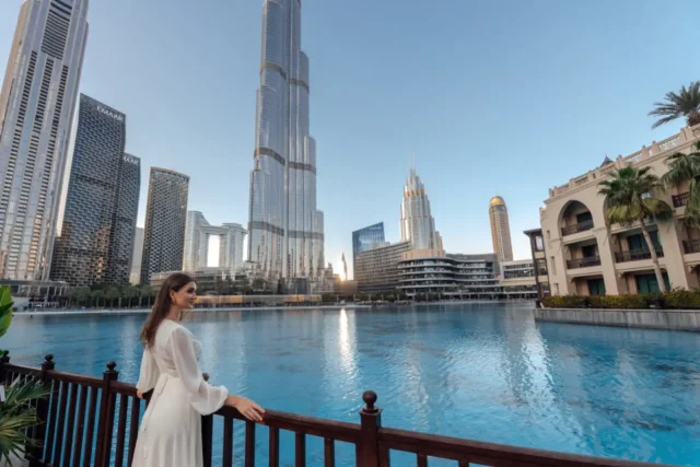 A woman in a white dress stands by a wooden railing overlooking a serene water body, with the Burj Khalifa and surrounding skyscrapers of Dubai in the background under a clear blue sky.